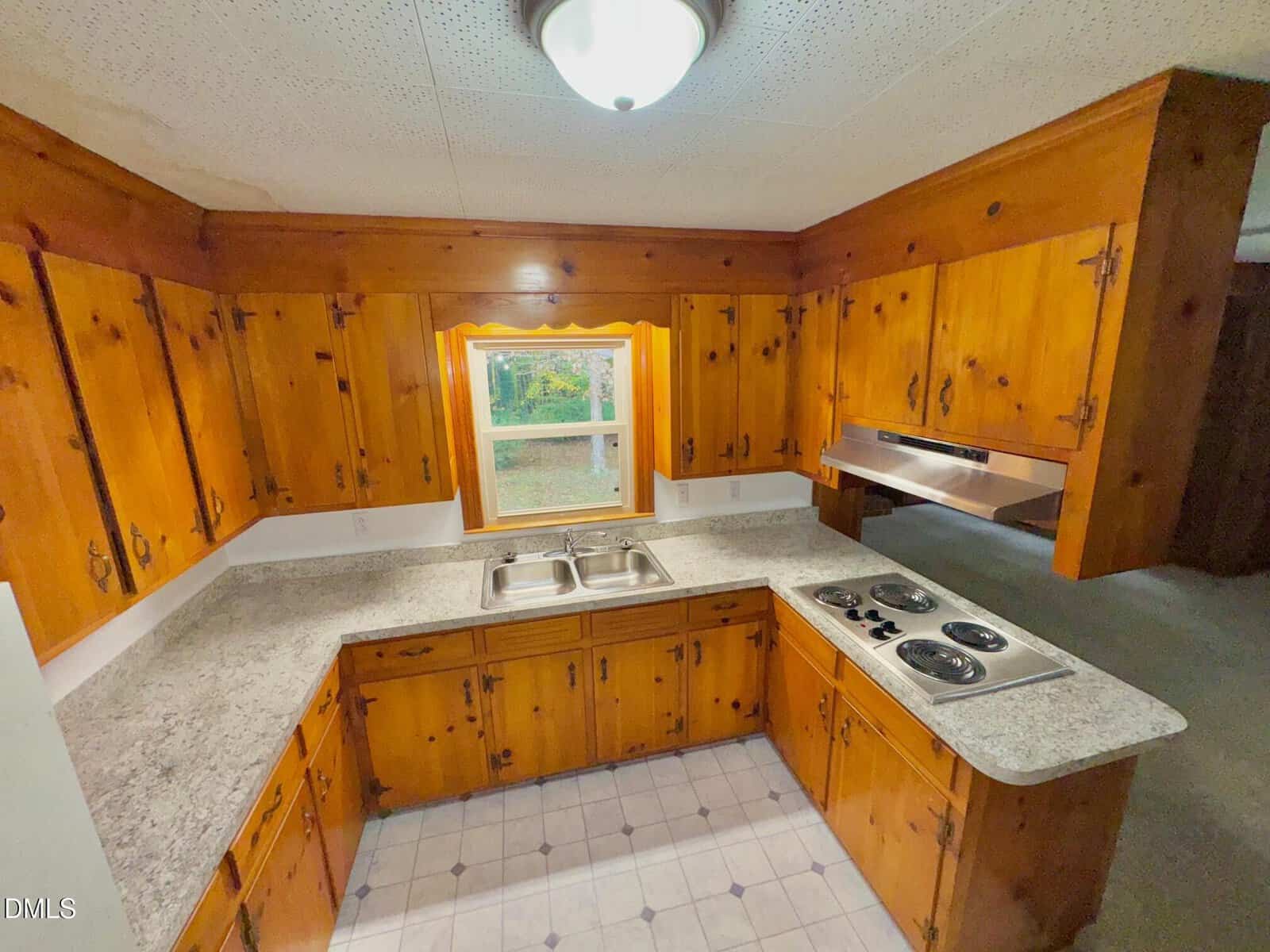A small kitchen with wooden cabinets, a double sink beneath a window, a four-burner electric stovetop, granite countertops, tile flooring, and a ceiling light fixture. A range hood is above the stovetop.