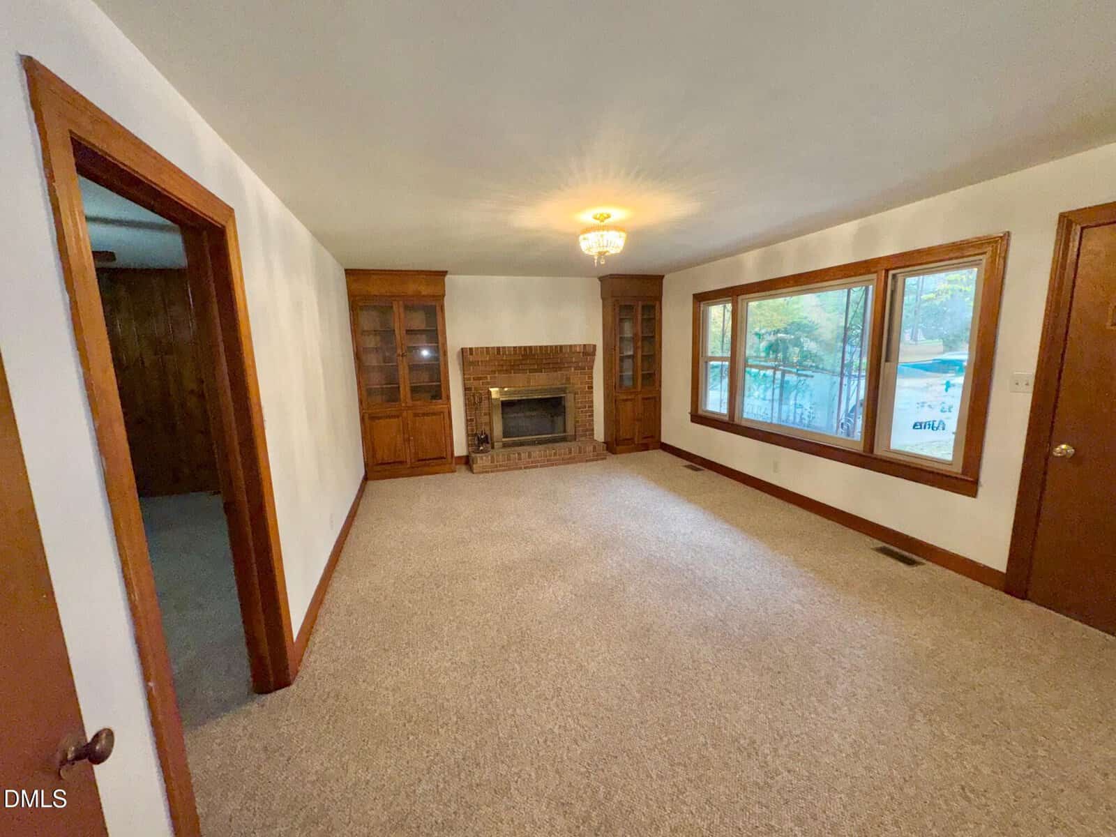 A carpeted living room with white walls, a brick fireplace centered between two wooden built-in cabinets, large windows on one wall, a ceiling light fixture, and a wooden door on the right.