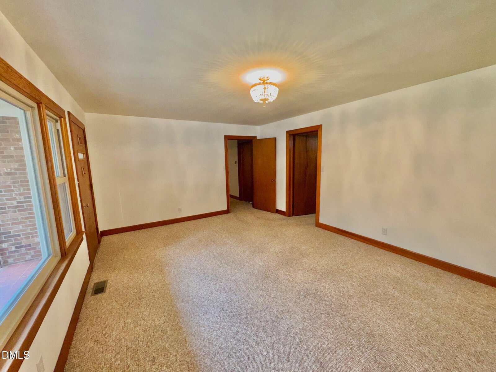 Unfurnished room with beige carpet, white walls, a ceiling light fixture, two wooden doors, a large window, and natural light coming through the window.