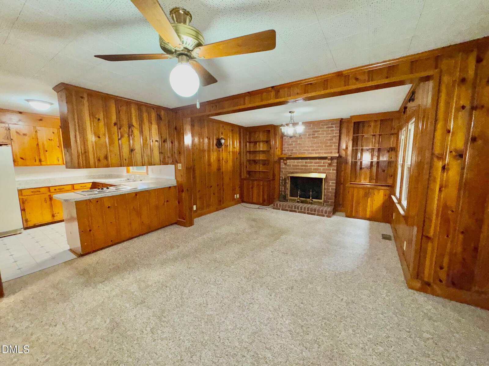 Room with wood-paneled walls, beige carpet, brick fireplace with built-in shelves, ceiling fan with light, and view of a kitchen area with white appliances and countertops.