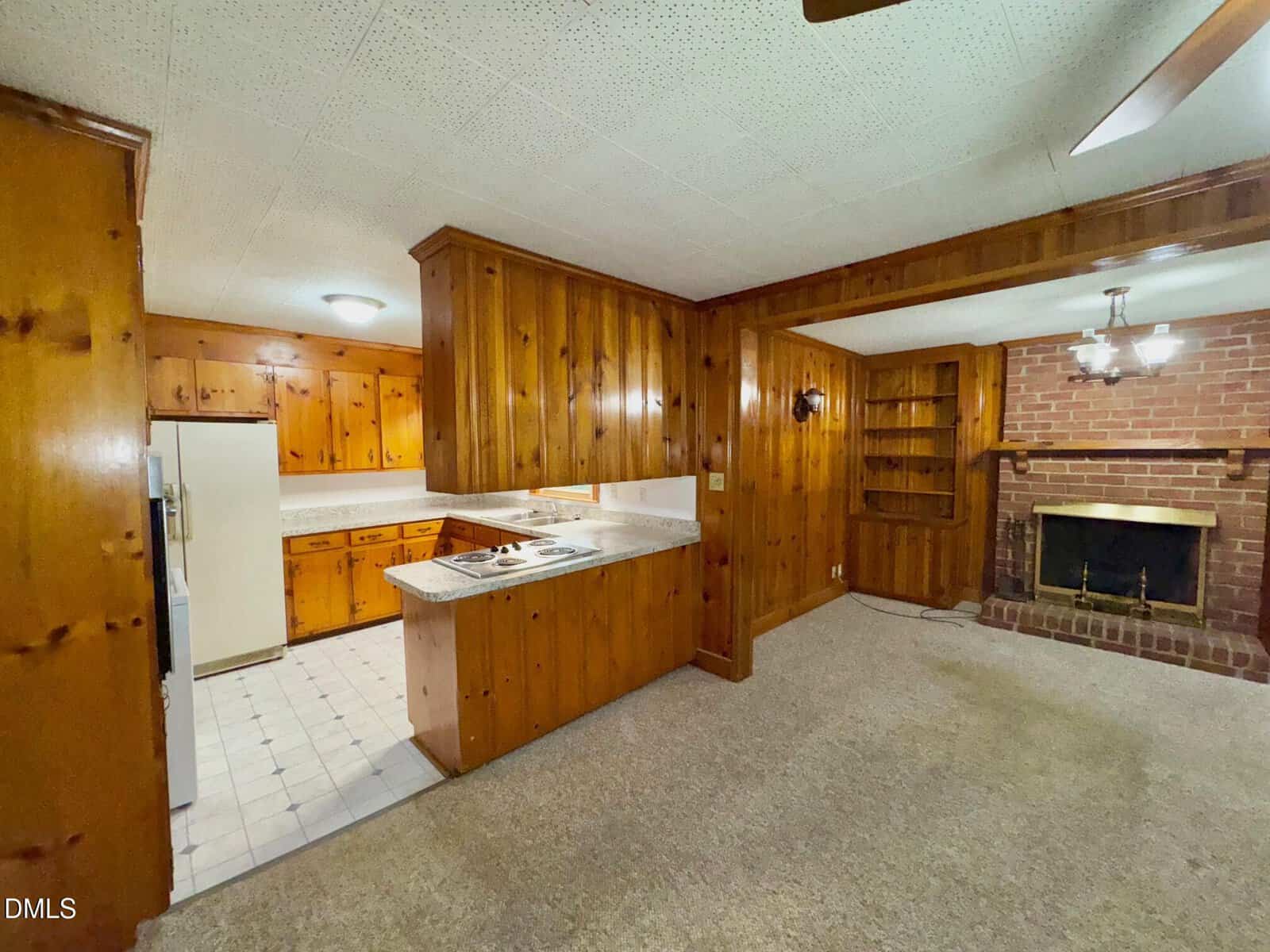 Wood-paneled room with carpeted floor, built-in shelves, and a brick fireplace. An open counter connects the space to a kitchen with white appliances and tile flooring. Ceiling lights illuminate both areas.