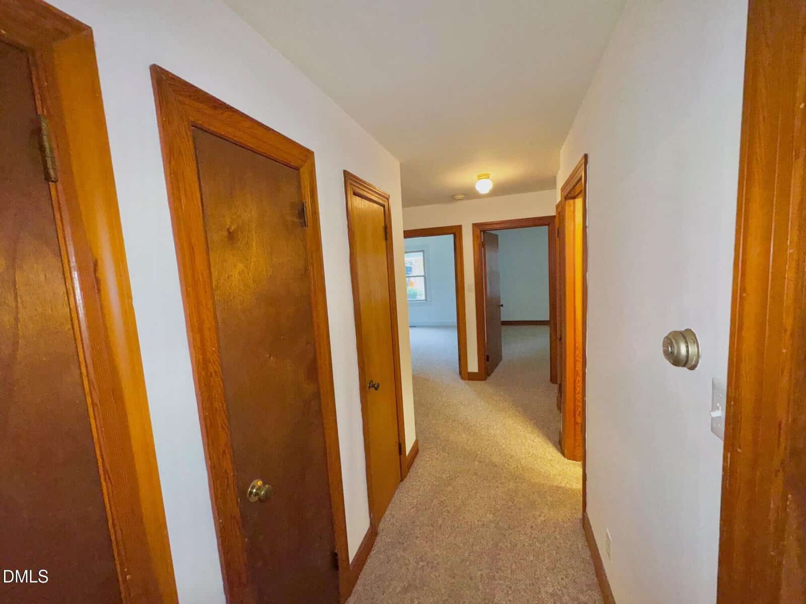 A hallway with beige carpet and white walls, featuring three wooden doors on the left and an open entryway leading to a bright room at the end. A light fixture is on the ceiling.
