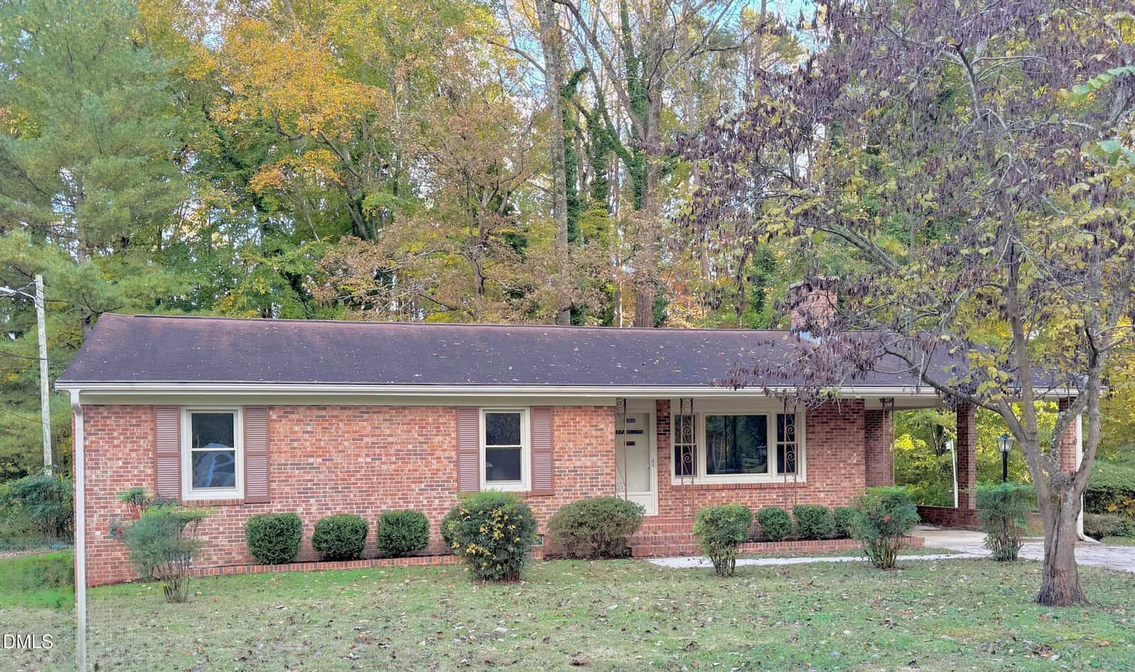 Charming single-story brick house at 105 Sunset Circle, Henderson, featuring a covered front porch, large front window, and dark shingle roof. Bushes line the home and a tree graces the lawn, with tall autumn trees in the background.