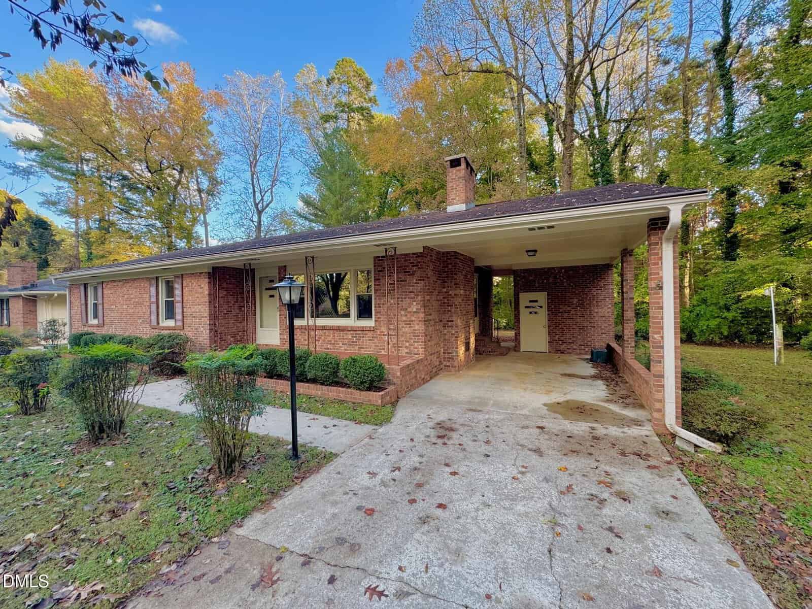 A single-story brick house with a carport on the right, a front yard with trimmed bushes and a lamp post, and trees with autumn leaves in the background. The driveway is concrete and partially covered.