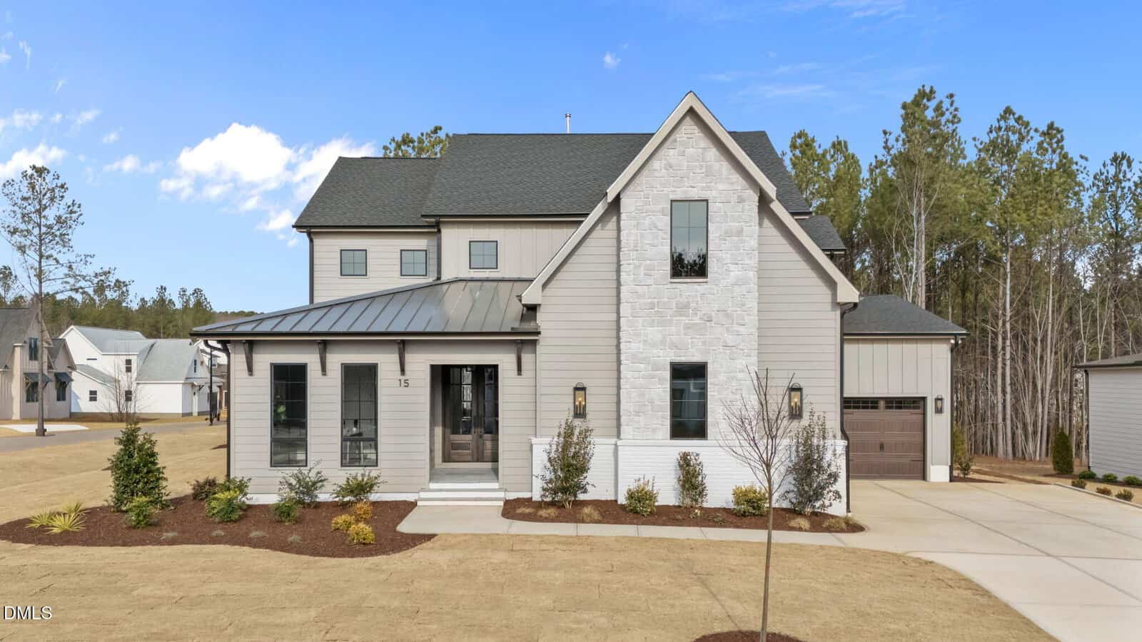 A modern two-story house with light gray siding, a dark roof, stone accents, and a single-car garage sits on a well-maintained lawn with small shrubs and trees. The sky is clear with some clouds.