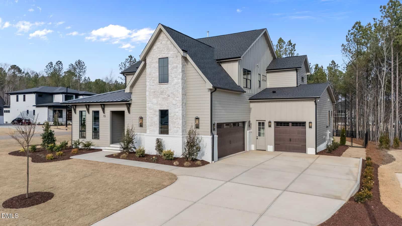 Two-story modern house with gray siding, white stone accent, and dark roof, featuring a three-car garage and a wide driveway. The lawn is landscaped with small shrubs and surrounded by trees.
