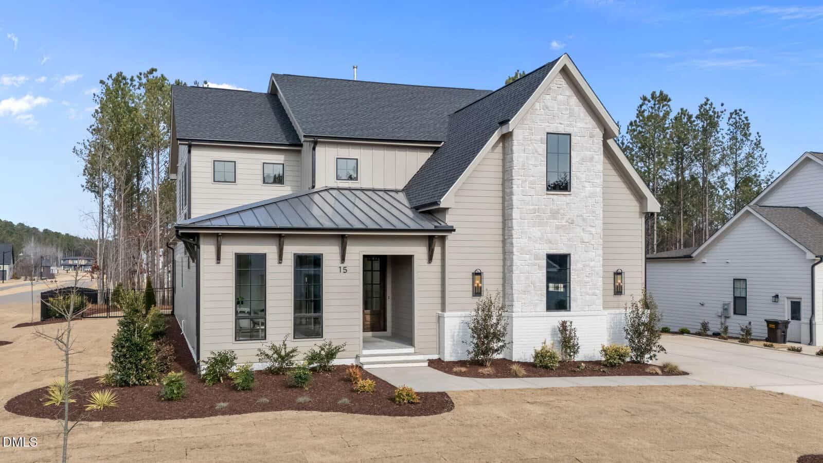 A two-story modern suburban house with gray siding, stone accents, black roof, and large windows. There are small shrubs and a mulched garden in front, with a neatly maintained lawn and a concrete walkway to the entrance.