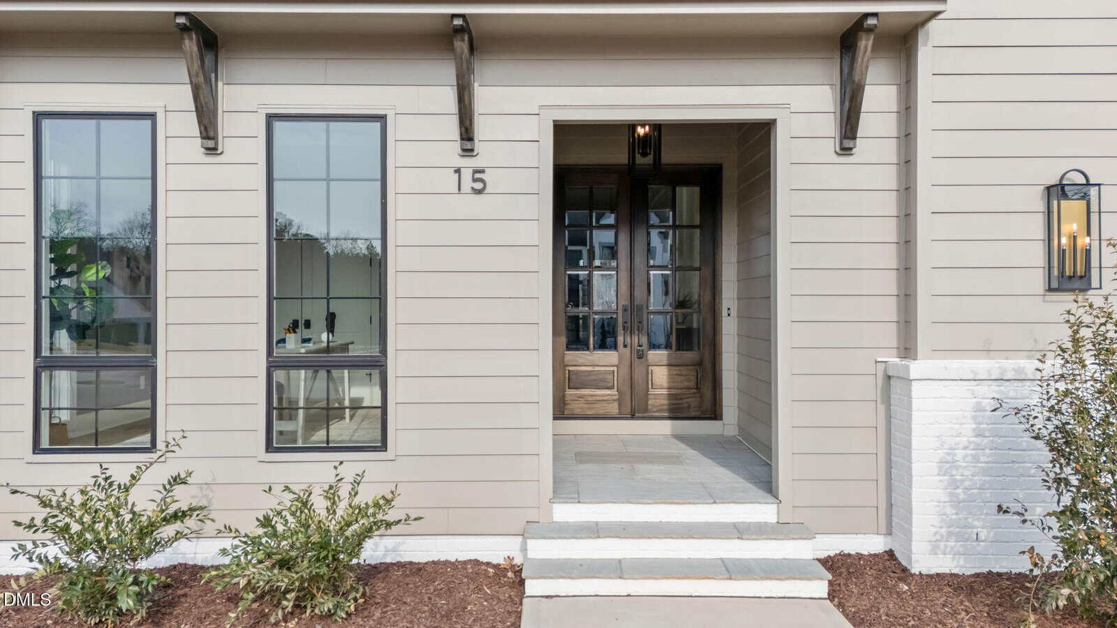 A modern house entrance with beige siding, large windows on either side of a wooden double front door, the number 15 displayed above the door, and shrubs planted along the walkway.