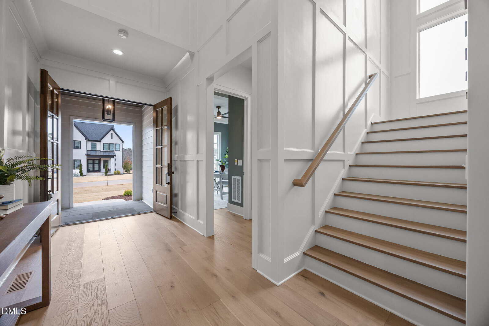 A spacious home entryway with light wood floors, white paneled walls, a wooden staircase with a light handrail, and a view through open double doors to a driveway and a modern house across the street.