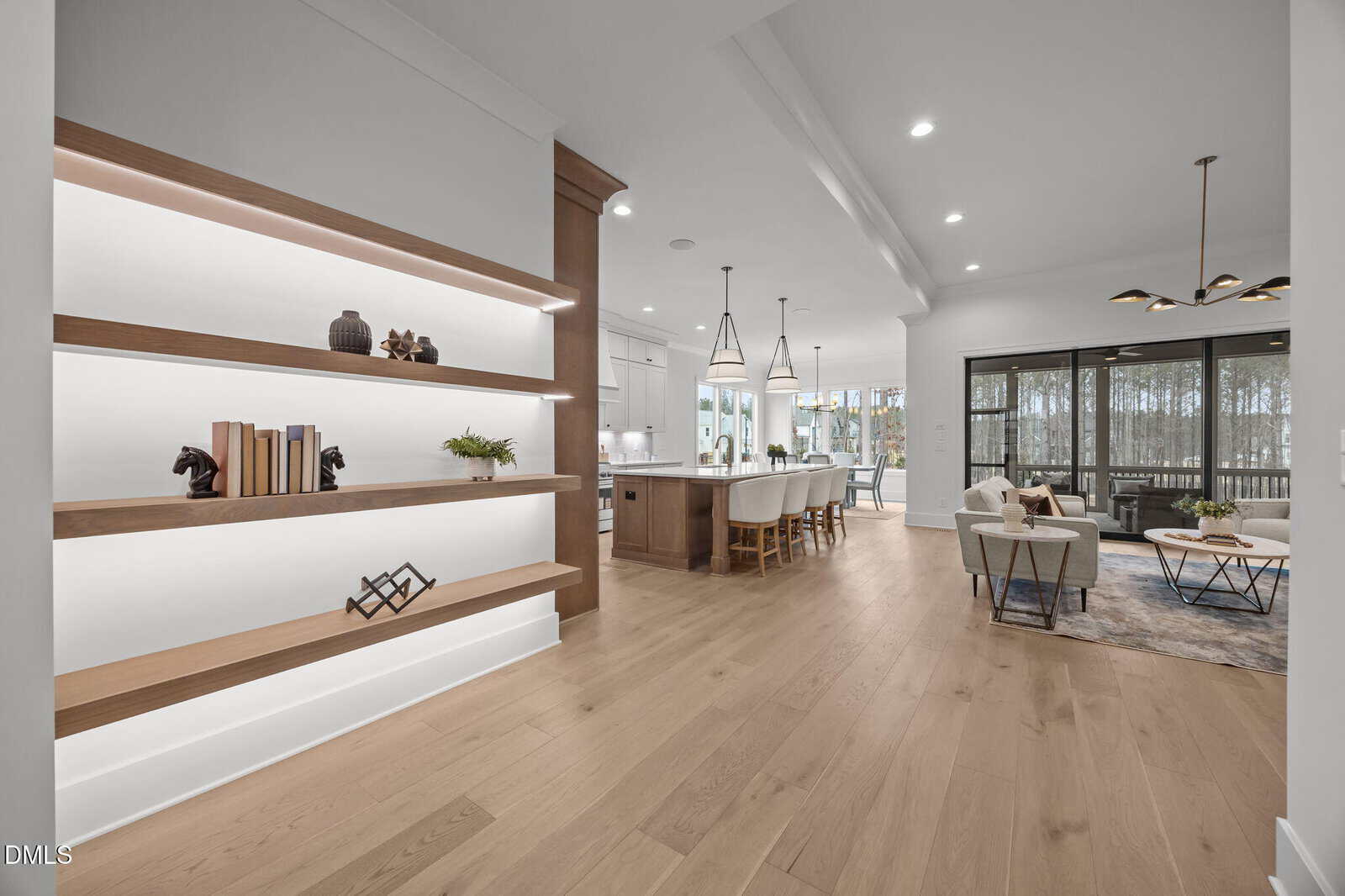 A modern open-concept living space with light wood floors, built-in shelves on the left, a kitchen with an island and stools in the center, and a seating area with large windows opening to an outdoor view.