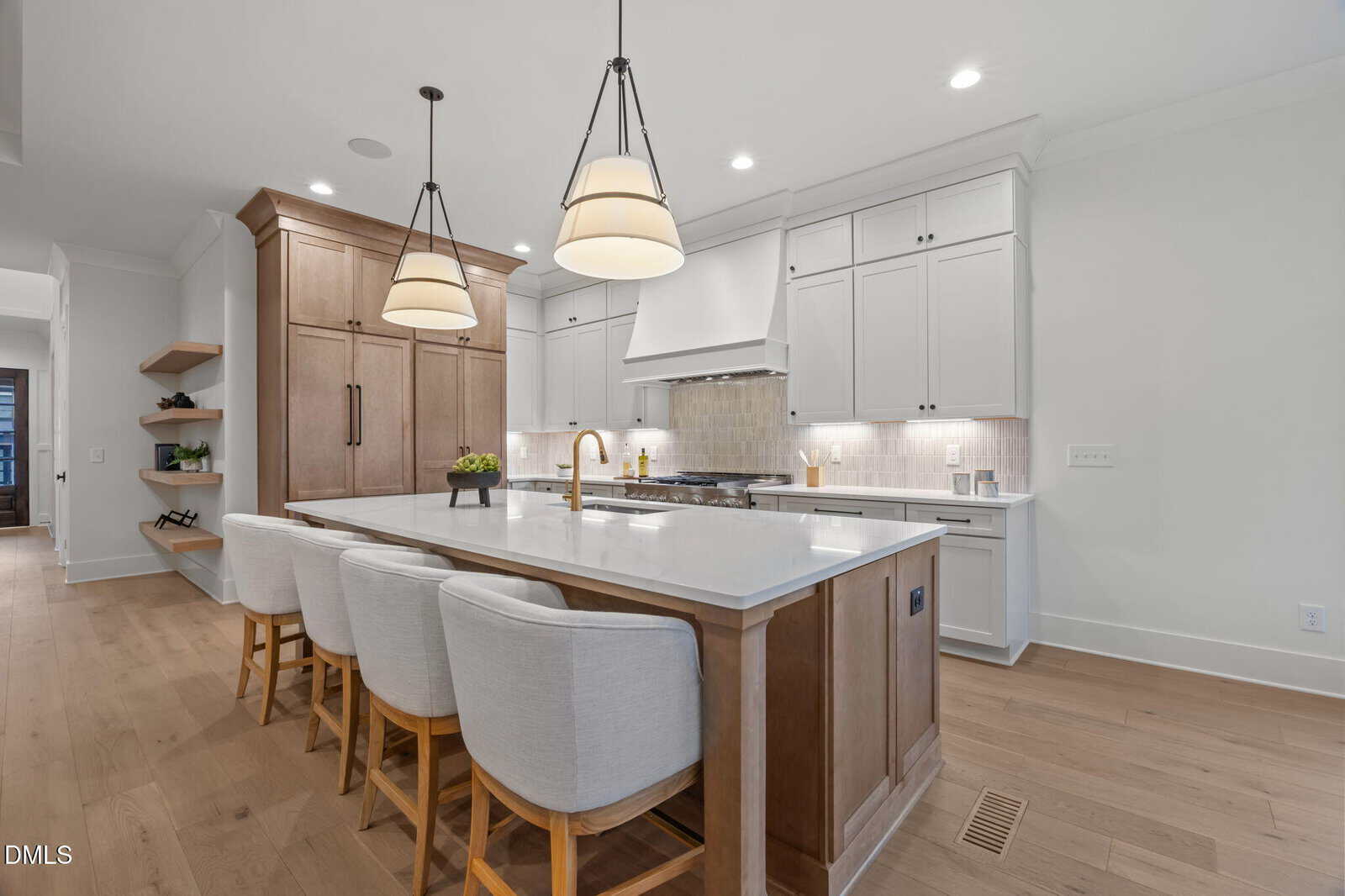 Modern kitchen with white cabinets, a large island with four upholstered chairs, pendant lights, wooden floors, and stainless steel appliances. Shelves and a built-in fridge are visible on the left.