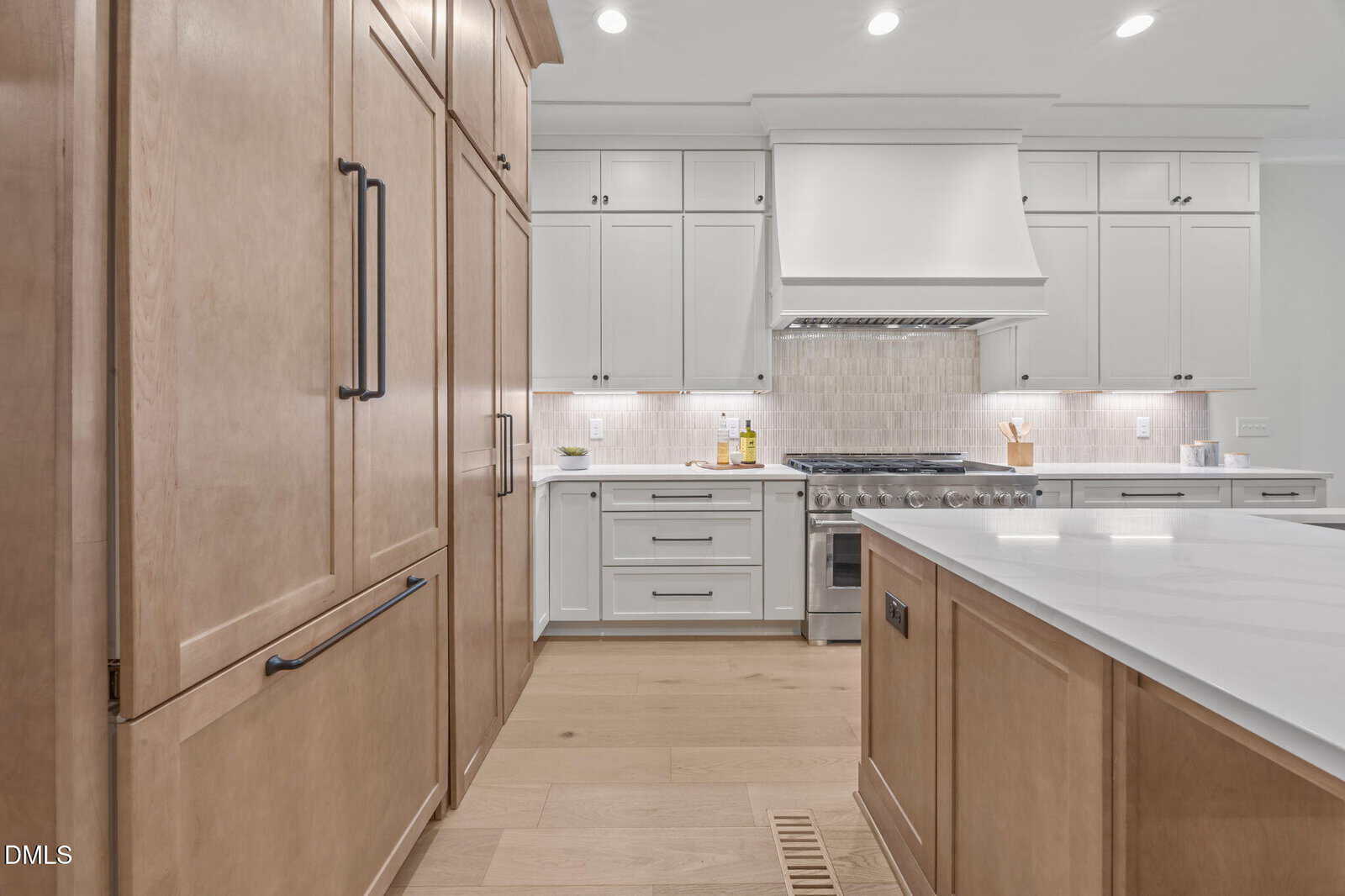 Modern kitchen with white cabinets, light wood floors, a large built-in fridge, stainless steel oven, white backsplash, and a spacious island with a white countertop.