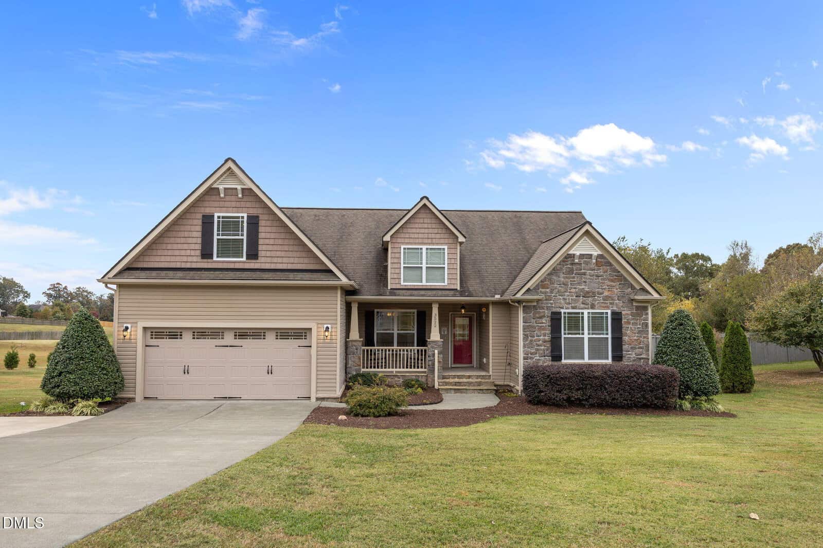 A suburban house in Wake Forest with a two-car garage, stone and siding exterior, covered front porch, and manicured lawn under a partly cloudy sky at 3522 Lavender Lane. Shrubs and trees are beautifully landscaped around the property.