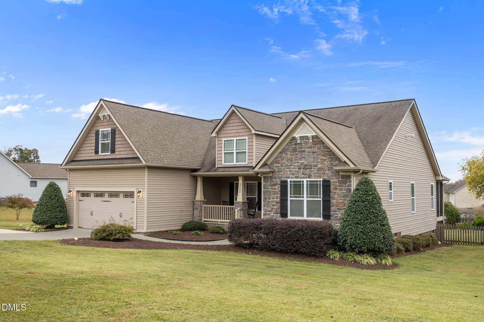Single-story suburban house with tan siding, a stone accent facade, and a two-car garage. There is a front porch, manicured bushes, and a large lawn under a blue sky with scattered clouds.