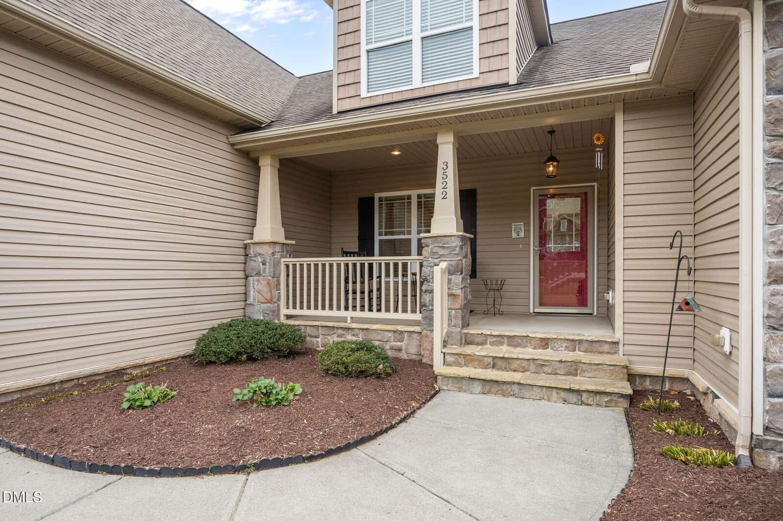 A front view of a house entrance with beige siding, stone accents, a covered porch with railing, and a small landscaped area with mulch and shrubs. The house number 3522 is displayed on a porch column.