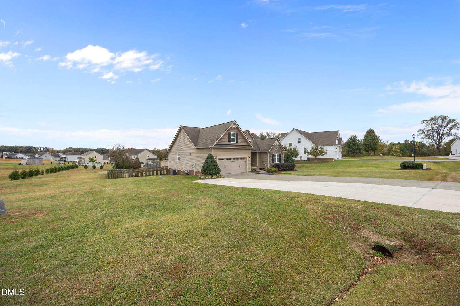 A tan two-story house with a two-car garage sits on a large, neatly mowed lawn. A wide concrete driveway leads to the garage, and other houses are visible in the background under a blue sky with clouds.