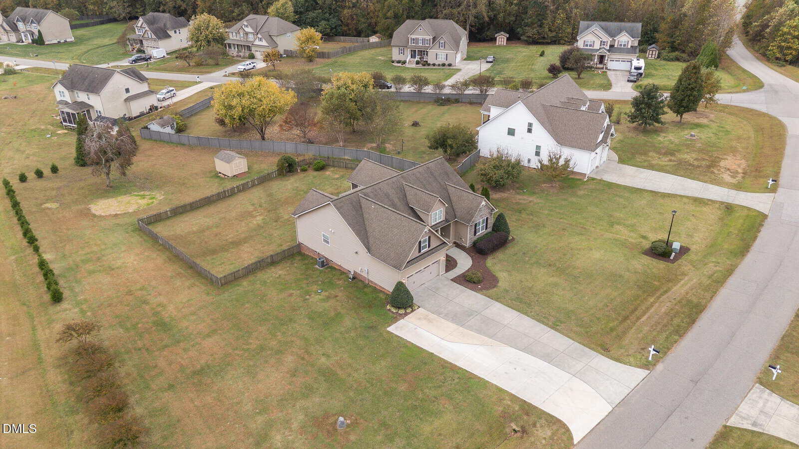 Aerial view of a suburban neighborhood with single-family homes, driveways, manicured lawns, fenced yards, trees, and a few sheds. The homes are spaced apart and bordered by roads and wooded areas.