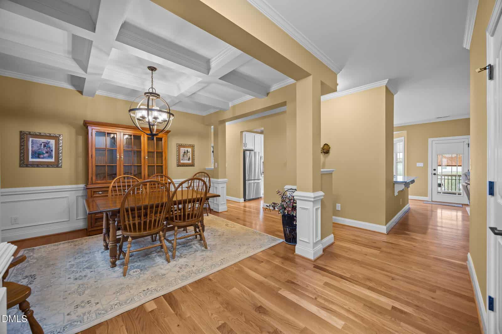 A dining room with a wooden table and six chairs sits on a rug under a coffered ceiling. A glass-front cabinet stands against the wall. The adjoining hallway leads to a kitchen and a door to the outside.