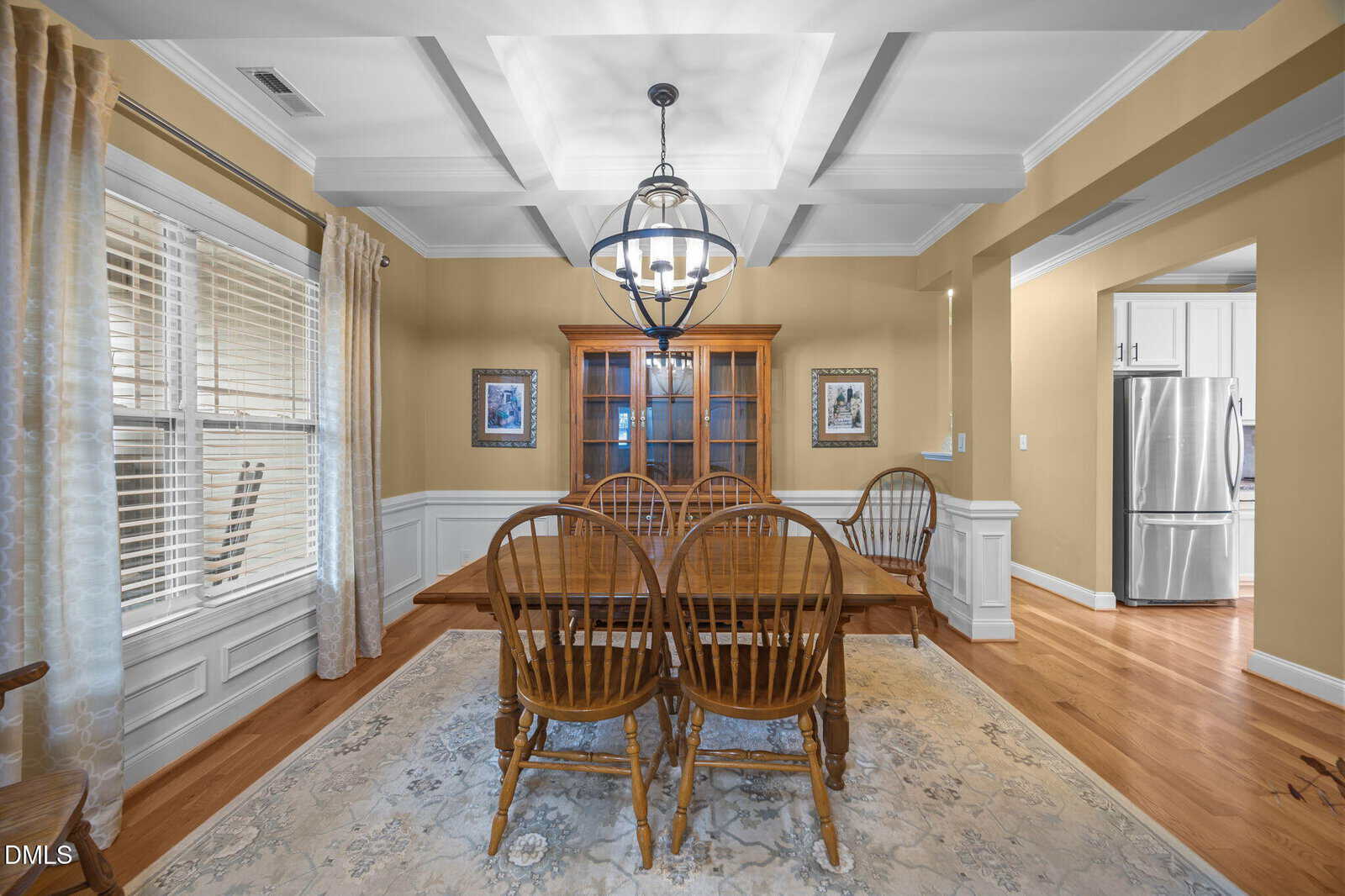 A formal dining room with a wooden table and six chairs, a light fixture above, a cabinet against the back wall, framed art, wood floors, and an open entryway to a kitchen with a stainless steel refrigerator.