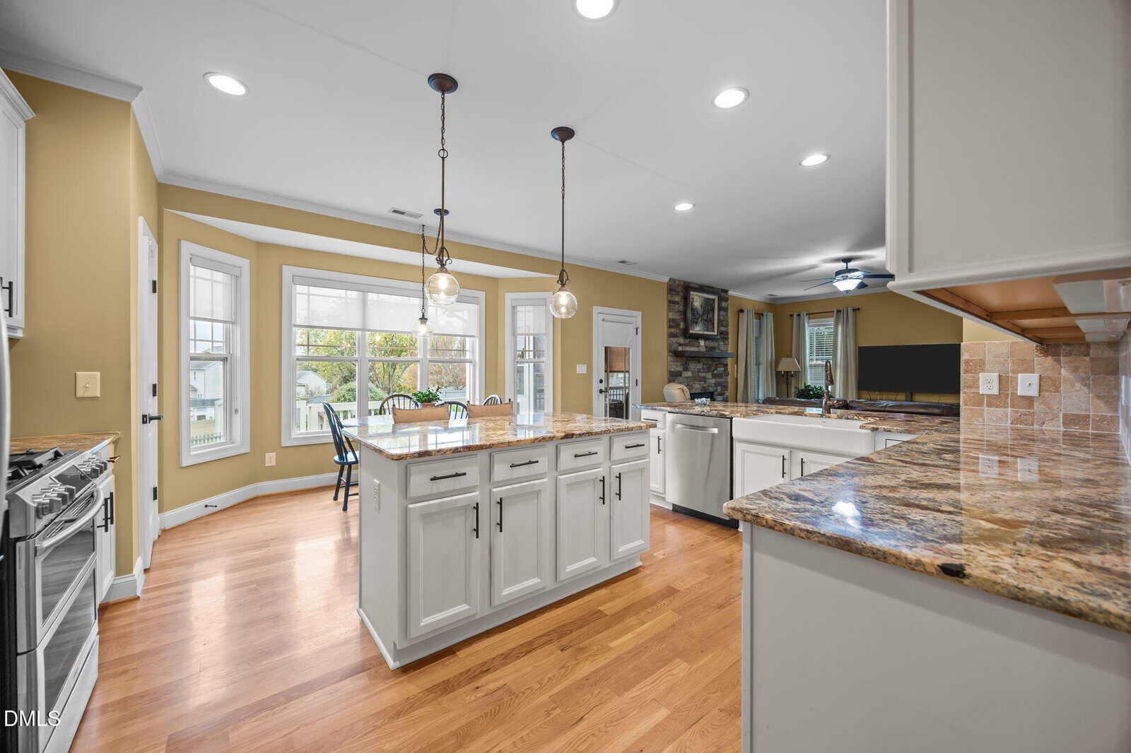 Spacious kitchen with white cabinets, granite countertops, a large island, hanging pendant lights, hardwood floors, and an open view to a dining area and living room with large windows.