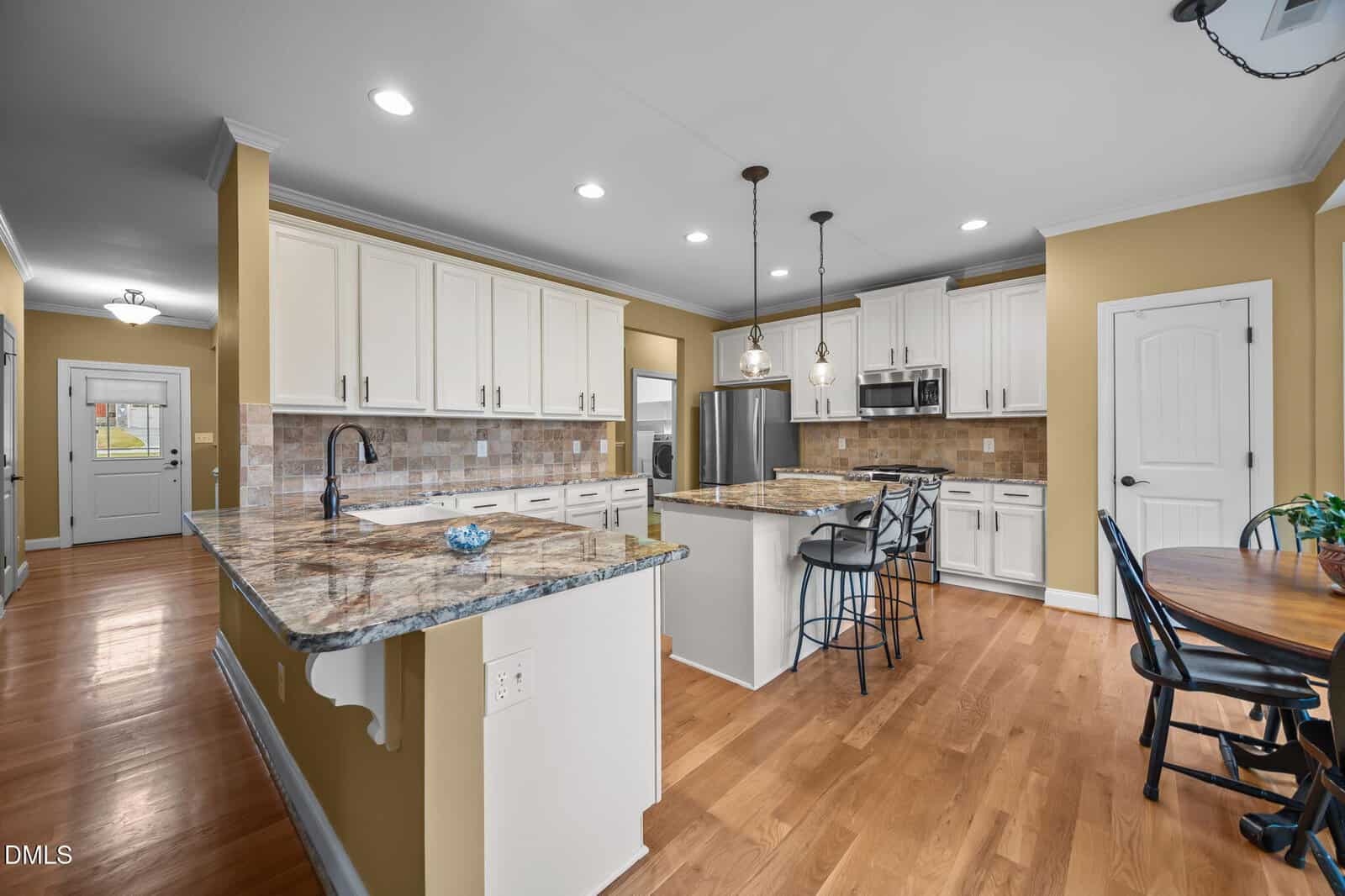 Modern kitchen with white cabinets, stainless steel appliances, granite countertops, a center island with barstools, pendant lighting, hardwood floors, and a small dining table with chairs near the window.
