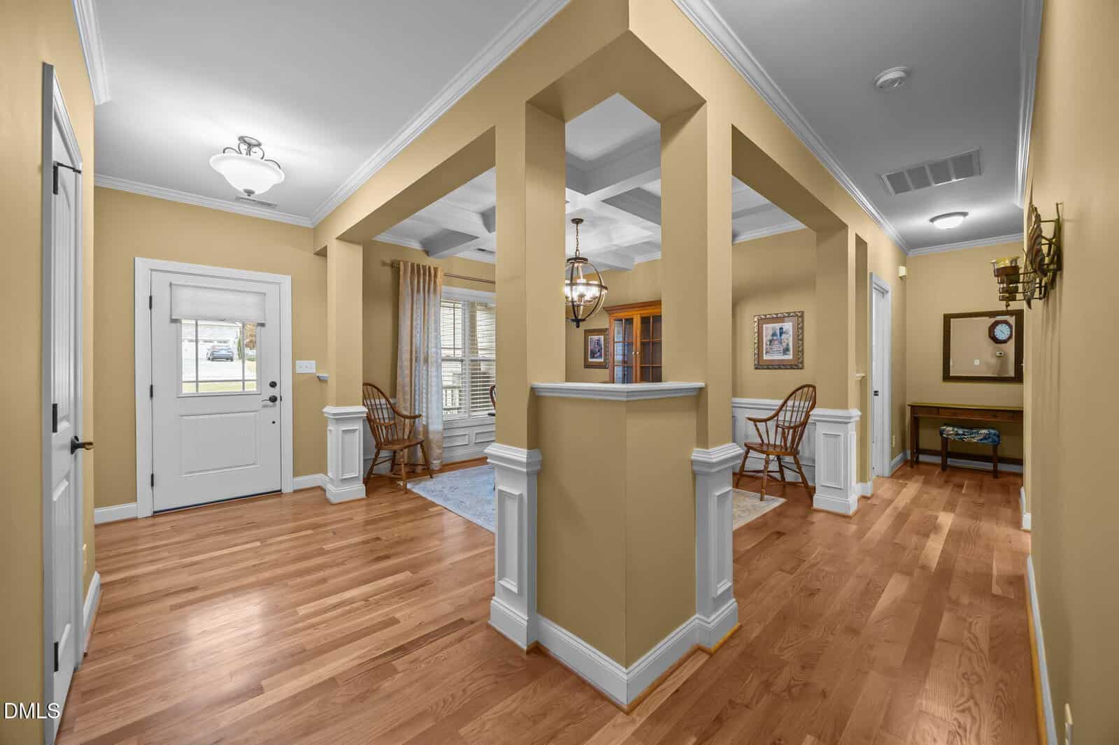 A hallway with light yellow walls, hardwood floors, white trim, and crown molding. The entry door is on the left, and wooden chairs, framed art, and a mirror decorate the space. Multiple doorways lead to other rooms.