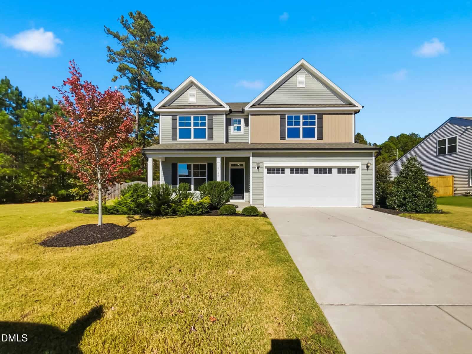 Two-story suburban house with gray siding, tan accent panel, white trim, and attached two-car garage. Neatly landscaped yard with a small tree, shrubs, and a concrete driveway. Blue sky and neighboring homes in view.