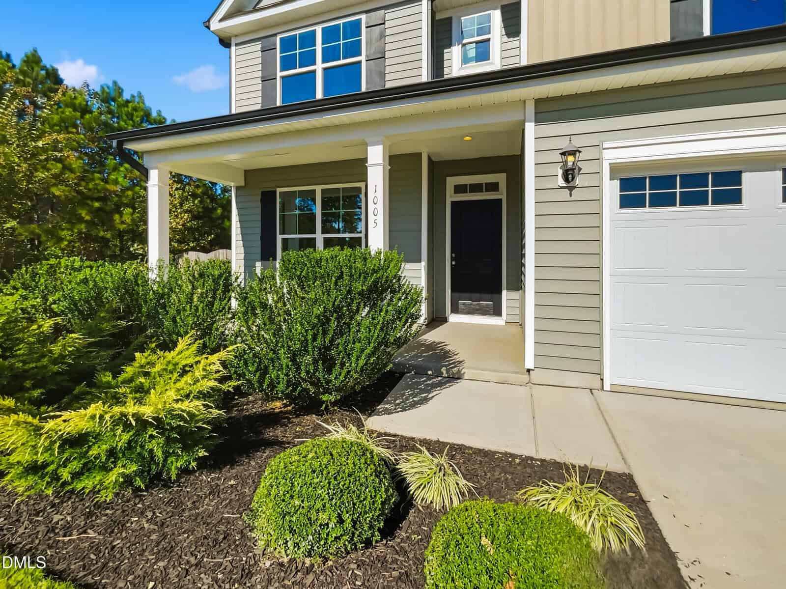 A two-story house with gray siding, white trim, a covered front porch, black front door, attached garage, and landscaped front yard with green bushes and mulch. The house number “1005” is visible on a porch column.