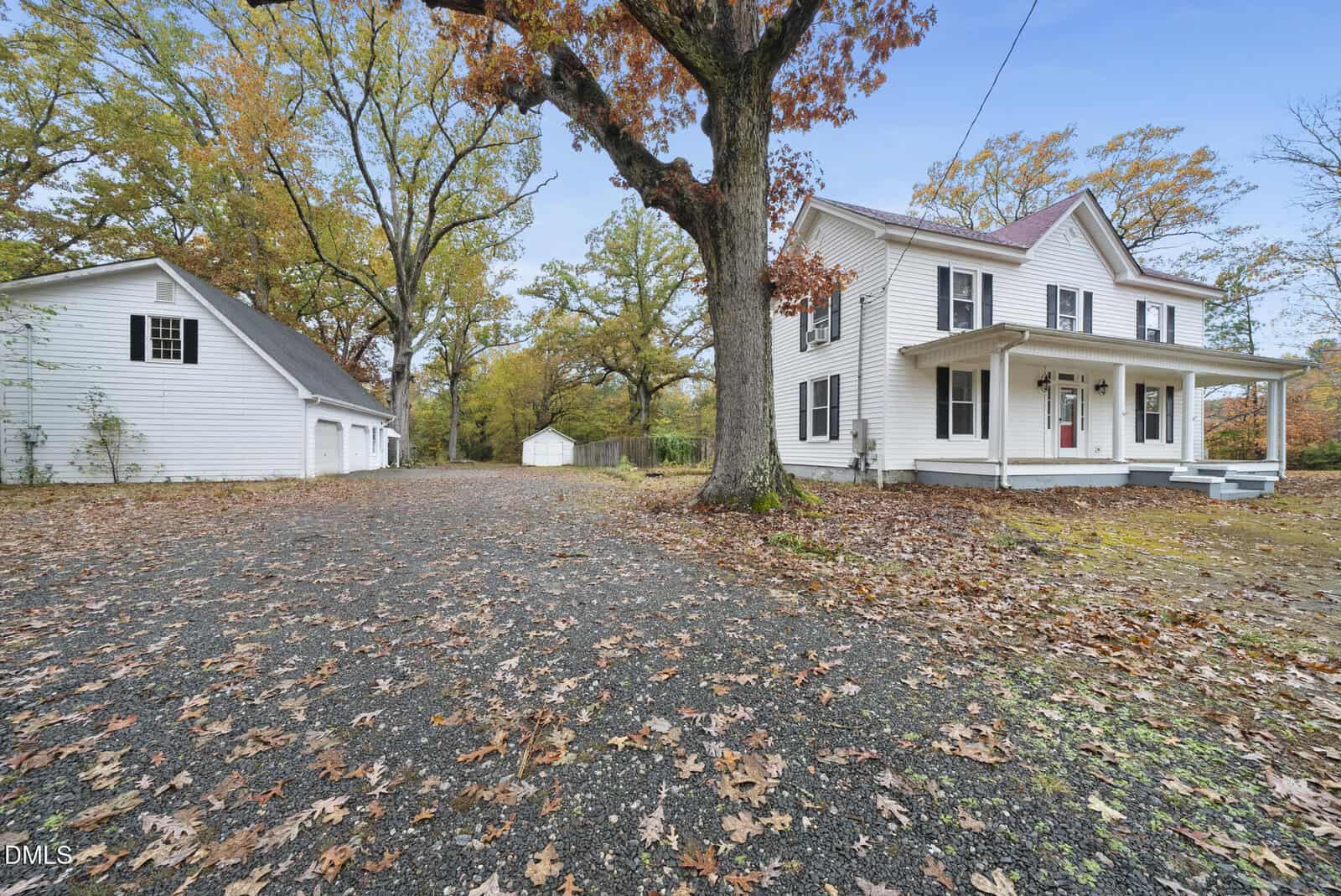 A white two-story house with a covered front porch sits beside a large tree on a leaf-covered driveway, with a detached white garage in the background, surrounded by autumn foliage.