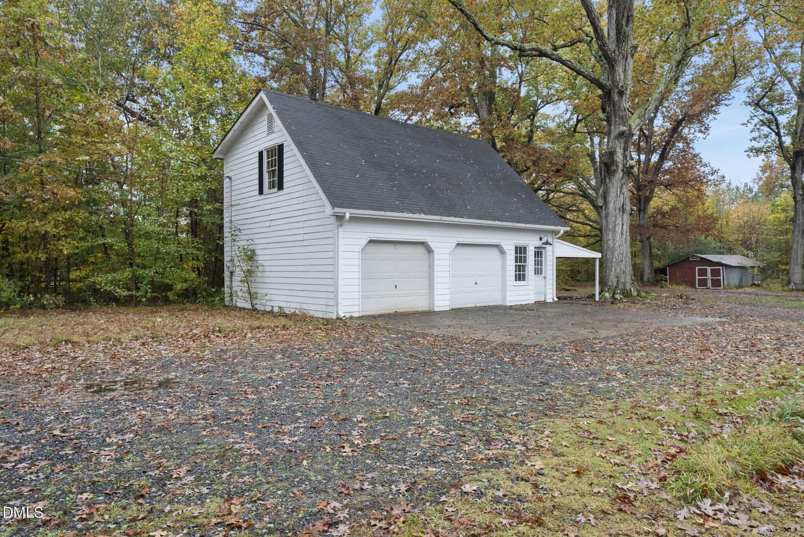 A white two-car garage with an upper story sits on a gravel driveway surrounded by trees with autumn leaves. A small red building is visible in the background among the trees.