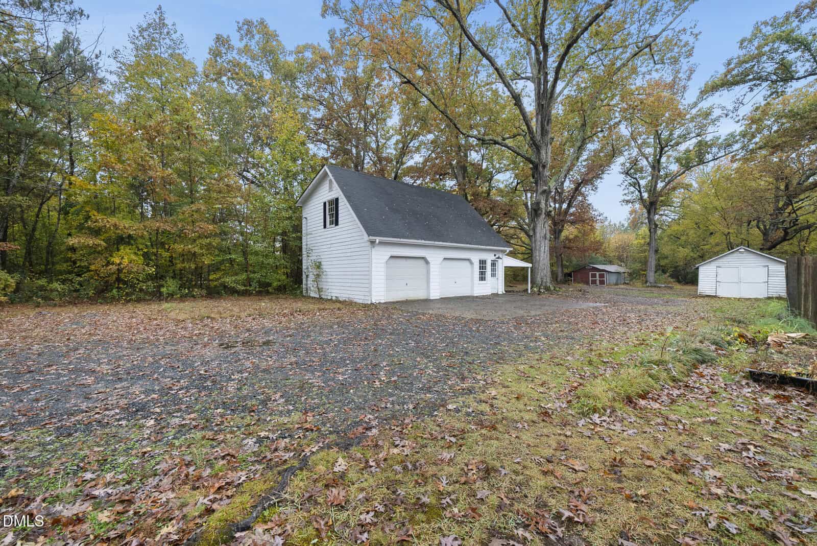 A white detached garage with two doors and an upper level sits in a wooded yard with scattered autumn leaves. A small shed and a red building are visible in the background among tall trees.