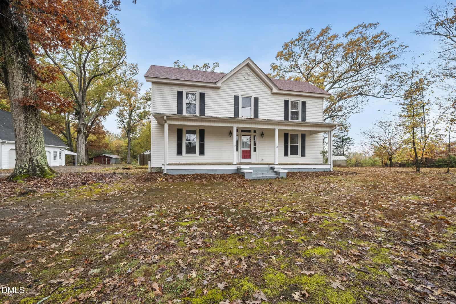 A white two-story house with a covered front porch, red front door, and black shutters sits on a large yard covered with fallen autumn leaves. The house is surrounded by tall trees.