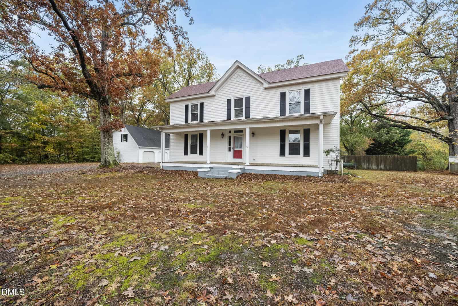 Two-story white house with black shutters, a covered front porch, and a red front door. Detached garage on the left, surrounded by trees with autumn leaves on the ground.
