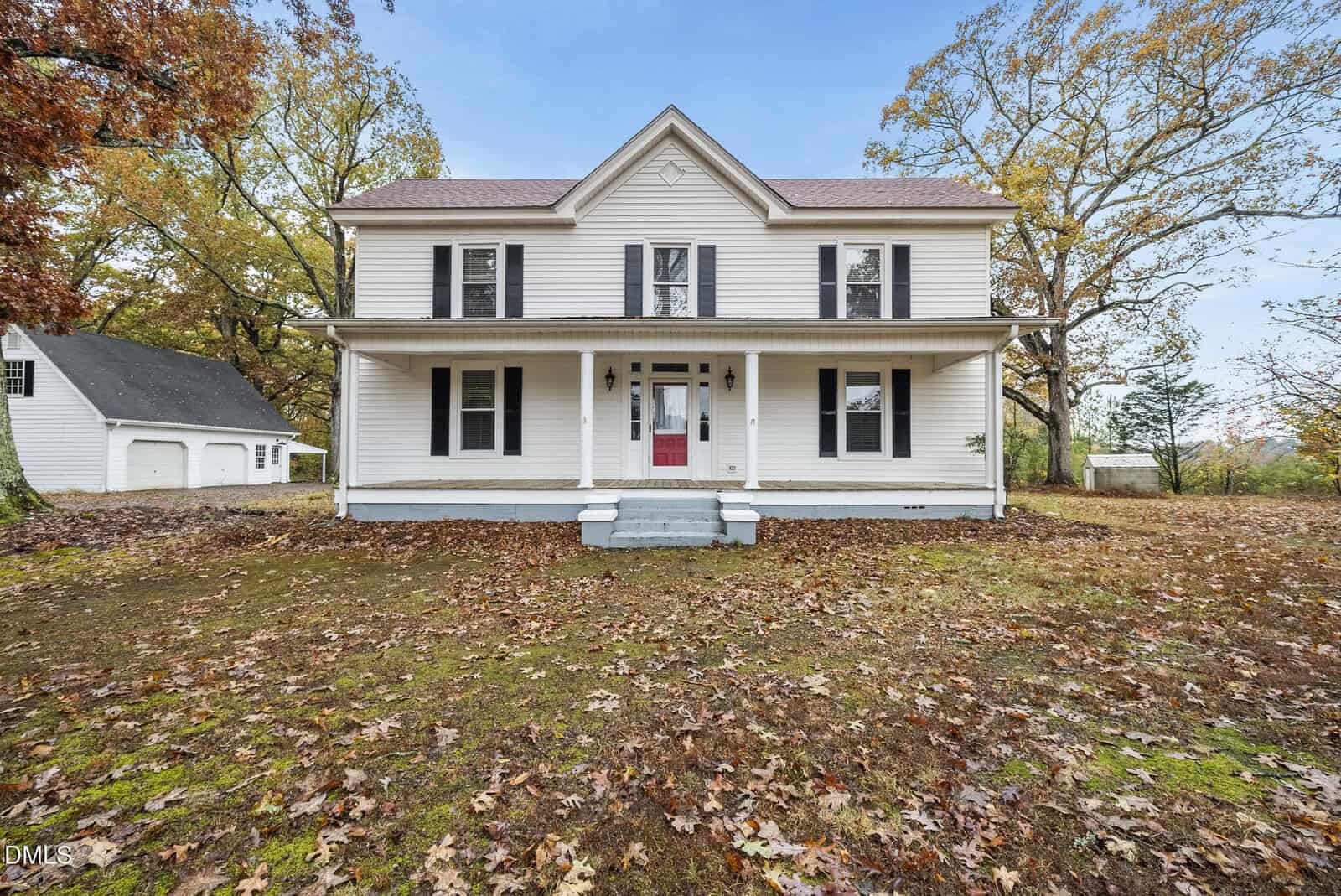 A charming two-story white house in Oxford with a covered front porch, black shutters, and a red door sits among autumn trees and fallen leaves. A detached white garage is visible to the left at 5011 NC 96.