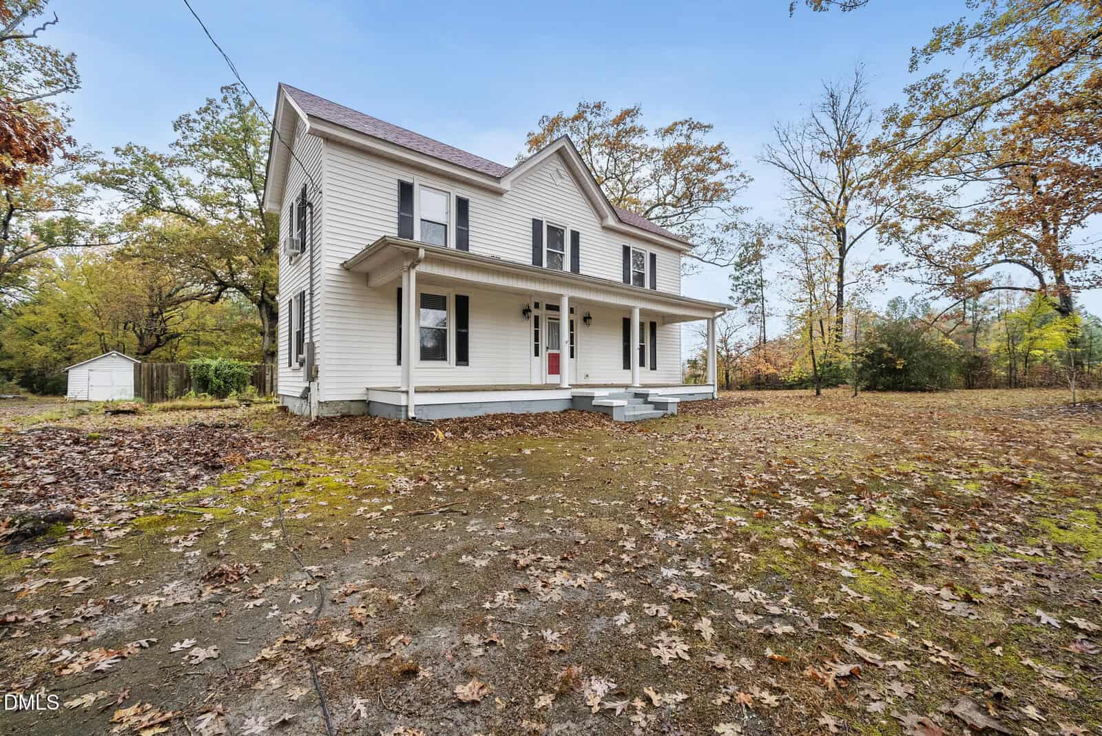 A two-story white house with black shutters and a covered front porch sits on a leaf-covered yard surrounded by trees. The sky is clear and there is a small white shed in the background.