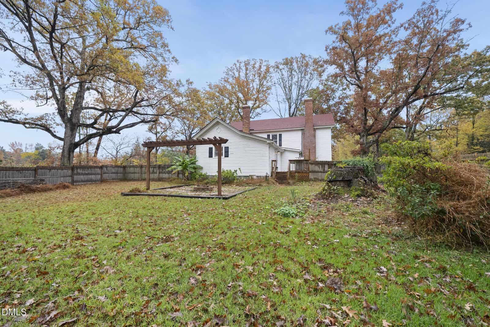 A white two-story house with a brown roof sits in a fenced yard. Trees with autumn leaves surround the property, and a wooden garden structure stands on the lawn.