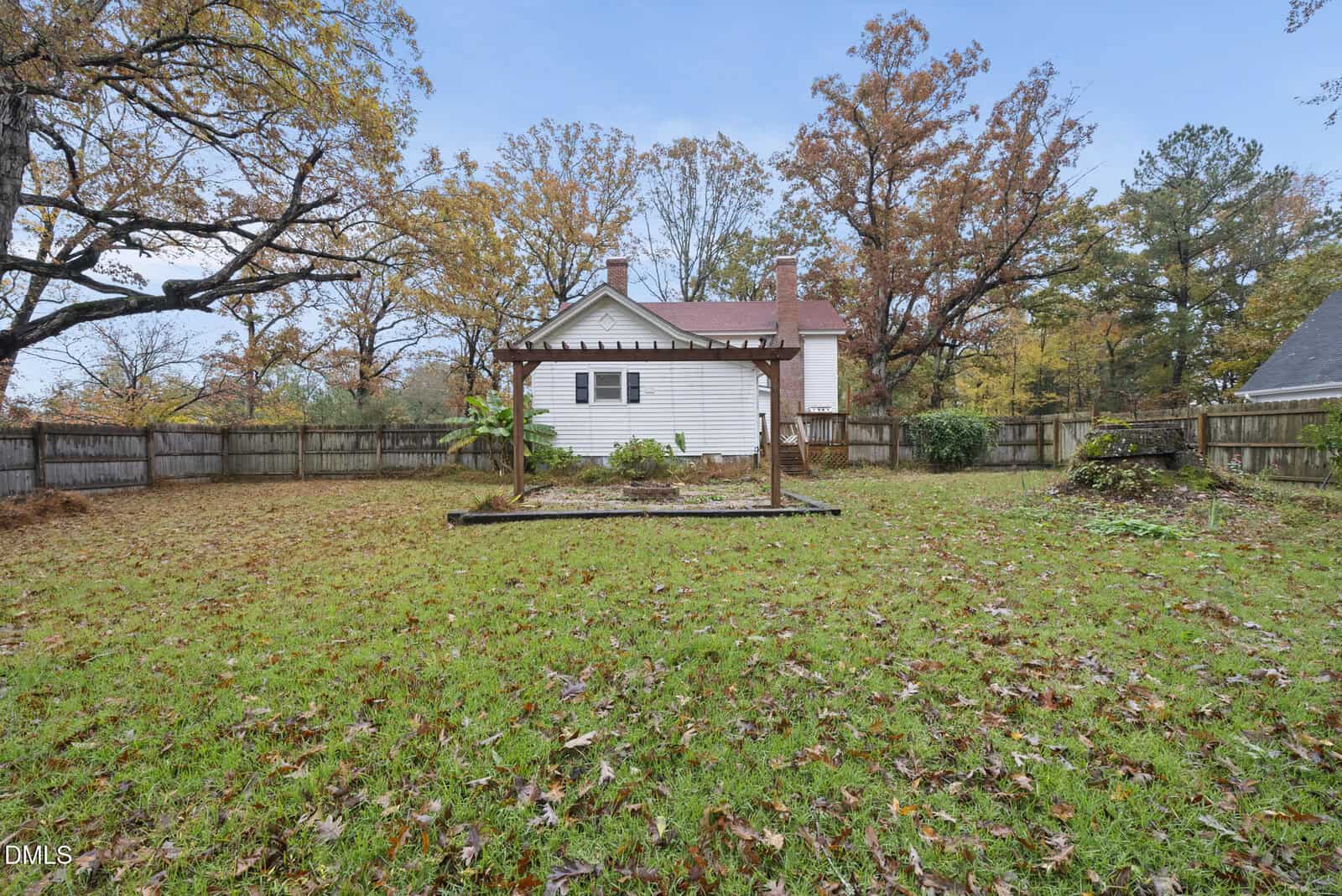 A backyard with a grassy lawn, wooden pergola, and a white house in the background. Fallen autumn leaves cover the ground, and tall trees with colored foliage surround the fenced yard.