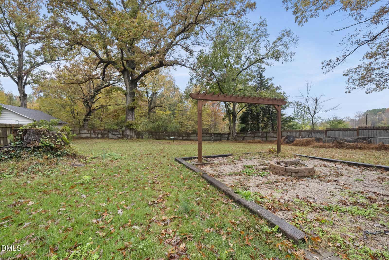 Spacious backyard with green grass, large trees, a wooden pergola, and a fire pit surrounded by gravel and wooden borders. Wooden fence in the background, leaves scattered on the ground, and a partly cloudy sky.