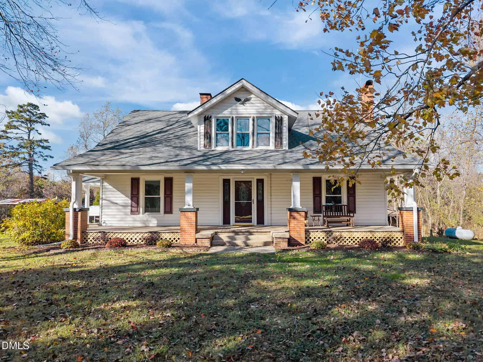 A white, two-story house at 595 George Sherman Road in Rougemont features a covered front porch with brick columns, a gabled roof, and a wooden porch swing. The grassy lot is dotted with trees, fallen leaves, and a propane tank in the background.