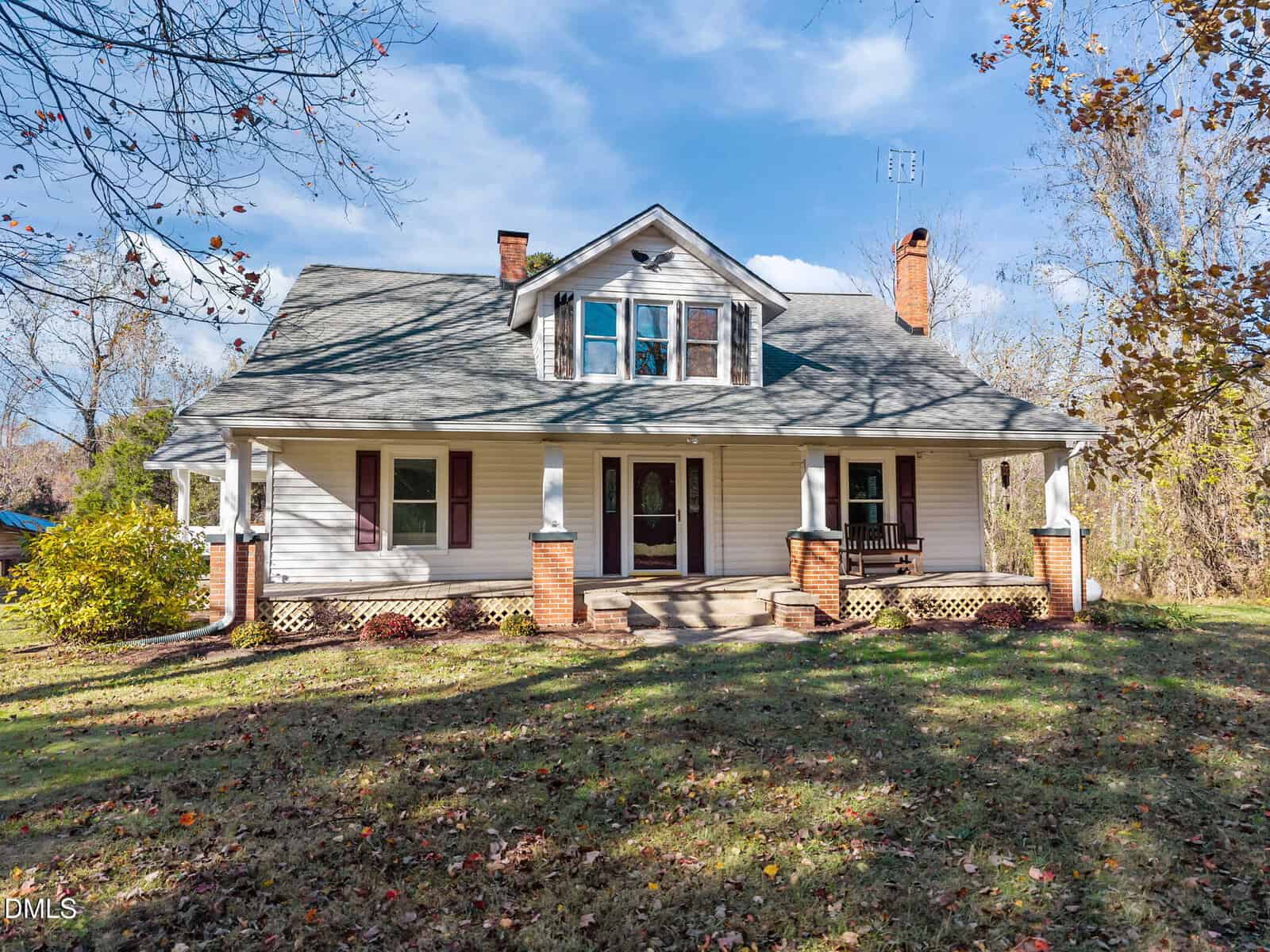 A white, two-story house with a gray roof, dormer windows, and two brick chimneys. The front porch features brick columns, a wooden swing, and steps leading to a grassy yard with scattered leaves. Trees surround the property.