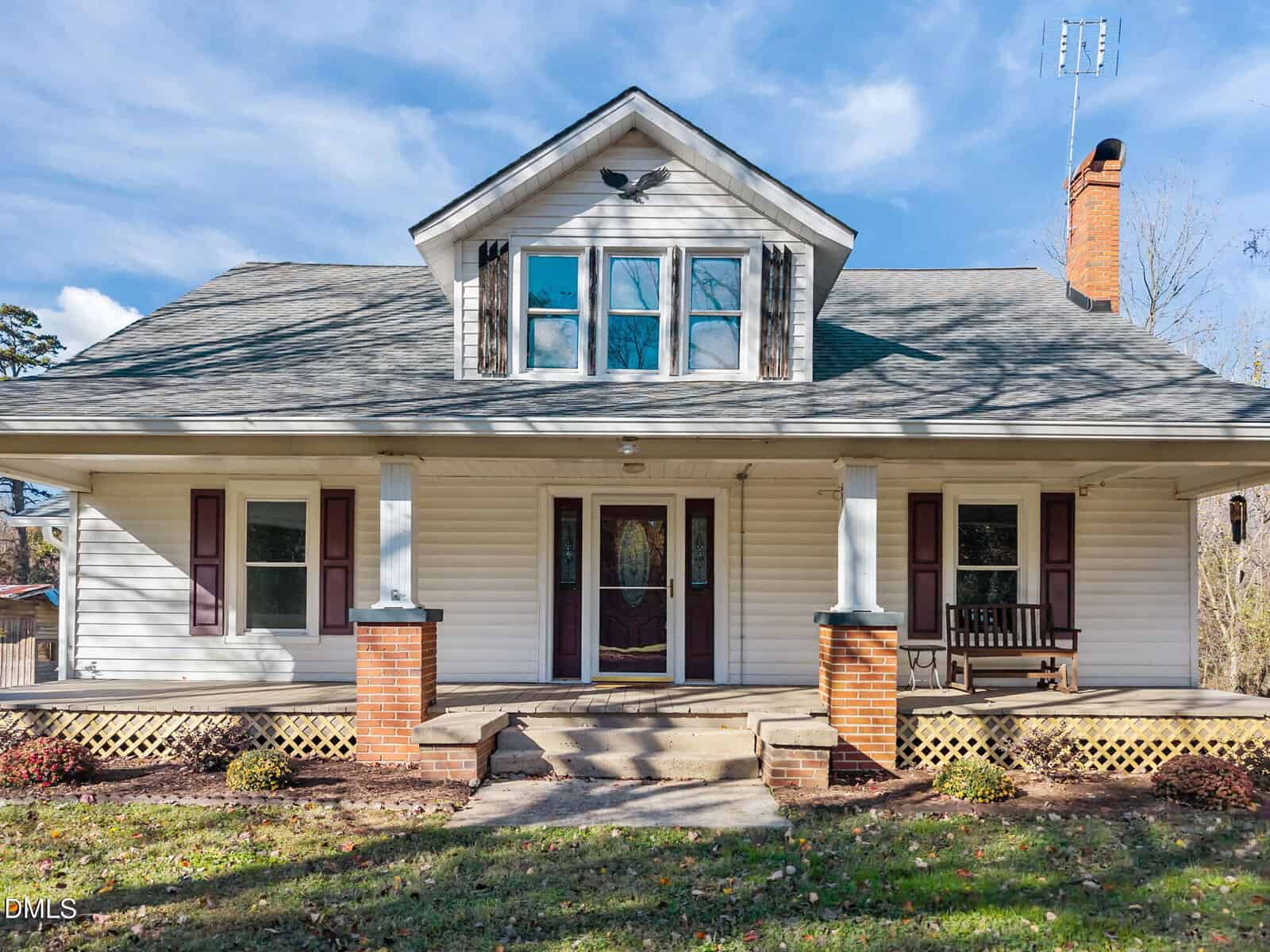 A white, single-story house with a covered front porch, brick columns, dark shutters, a dormer window, a wooden bench, and a chimney. The yard has minimal landscaping, and the sky is partly cloudy.
