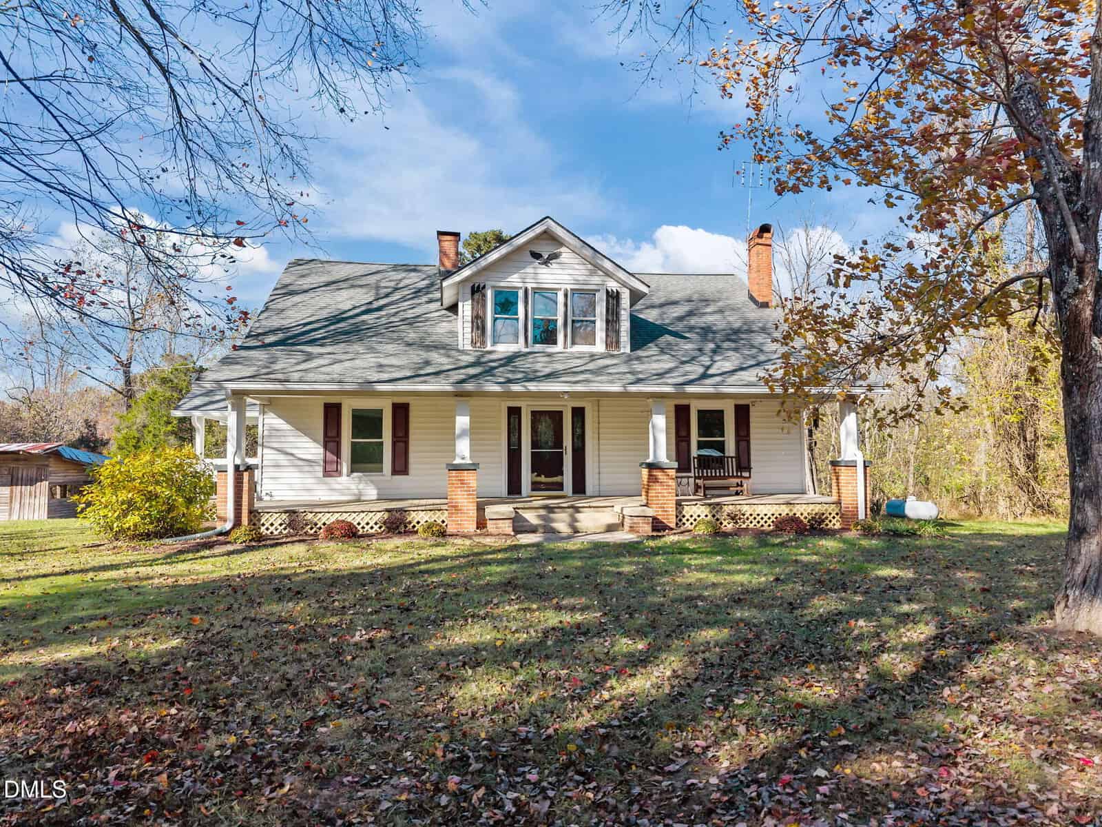A two-story white house with a covered front porch, brick columns, dormer windows, and a gabled roof, surrounded by a grassy yard with autumn leaves and trees.