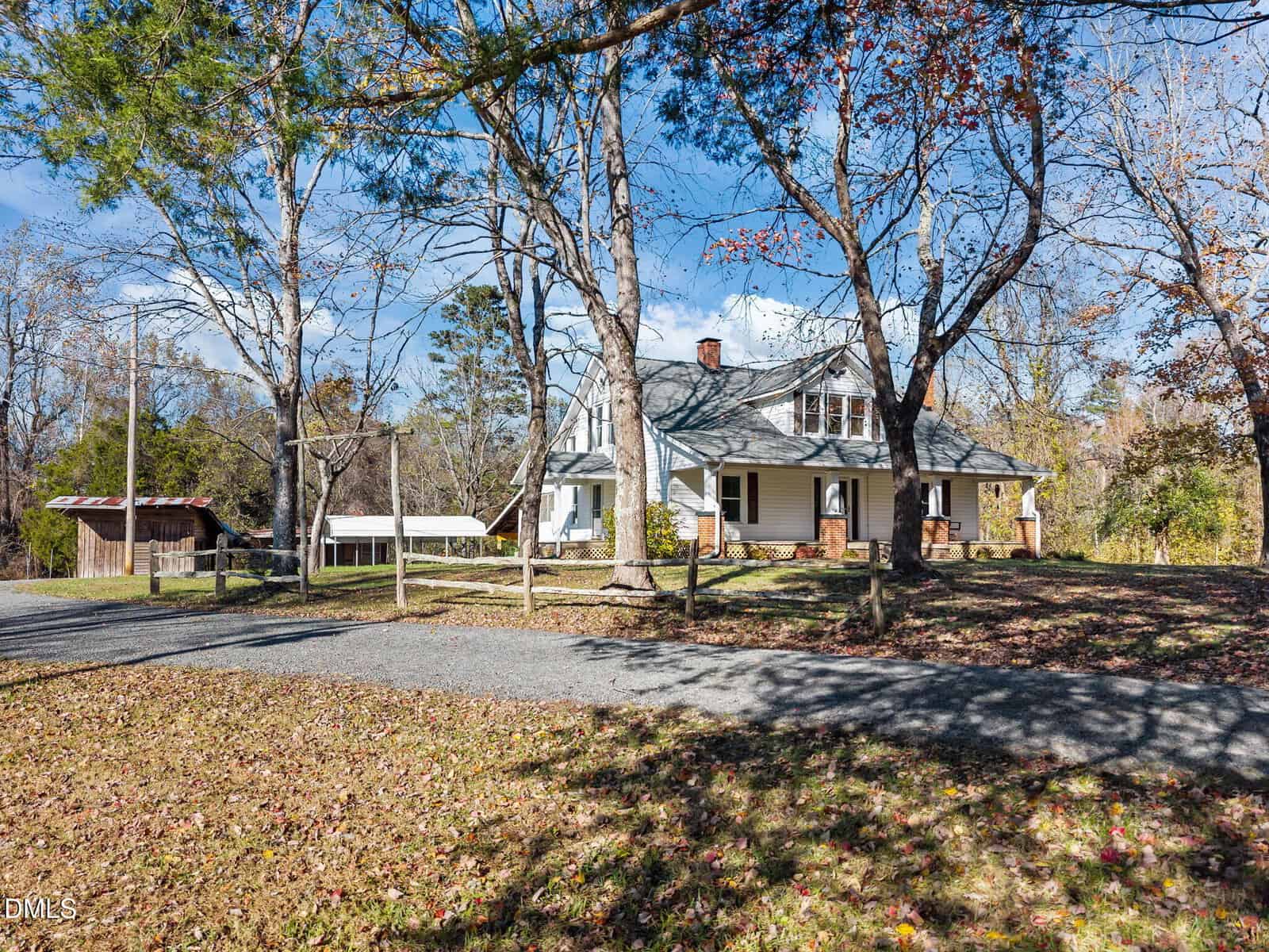 A two-story white house with a front porch sits among tall trees on a sunny day. A gravel driveway curves in front, and a wooden shed is visible to the left near the edge of the property.