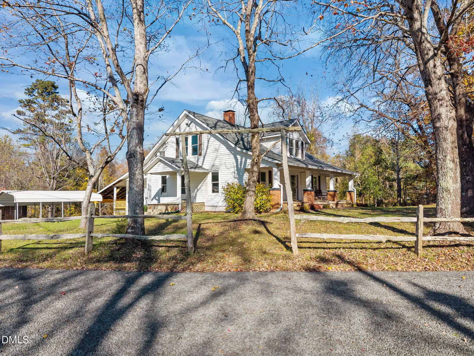 White two-story house with a covered front porch, set behind a wooden fence and trees with few leaves. The yard has grass and some shrubs. A driveway and detached carport are visible on the left side.