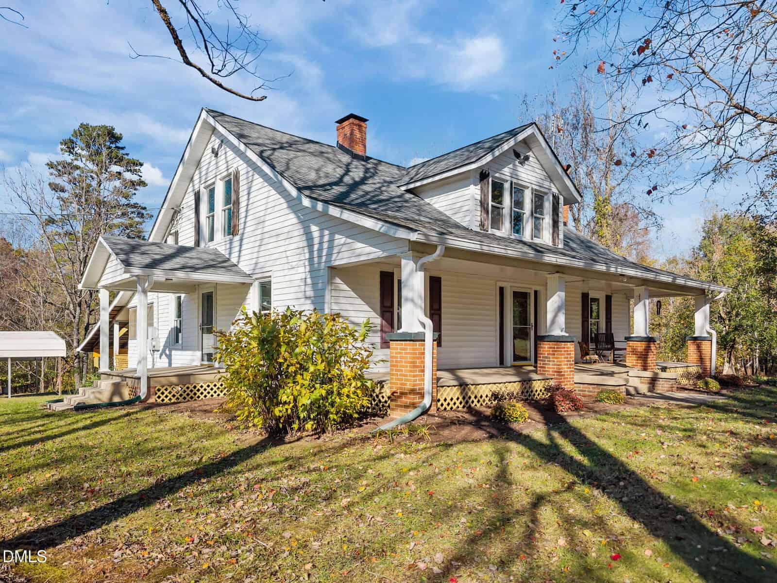 A white two-story house with a covered front porch supported by brick columns, surrounded by a grassy yard and trees with some fall foliage, under a clear blue sky.