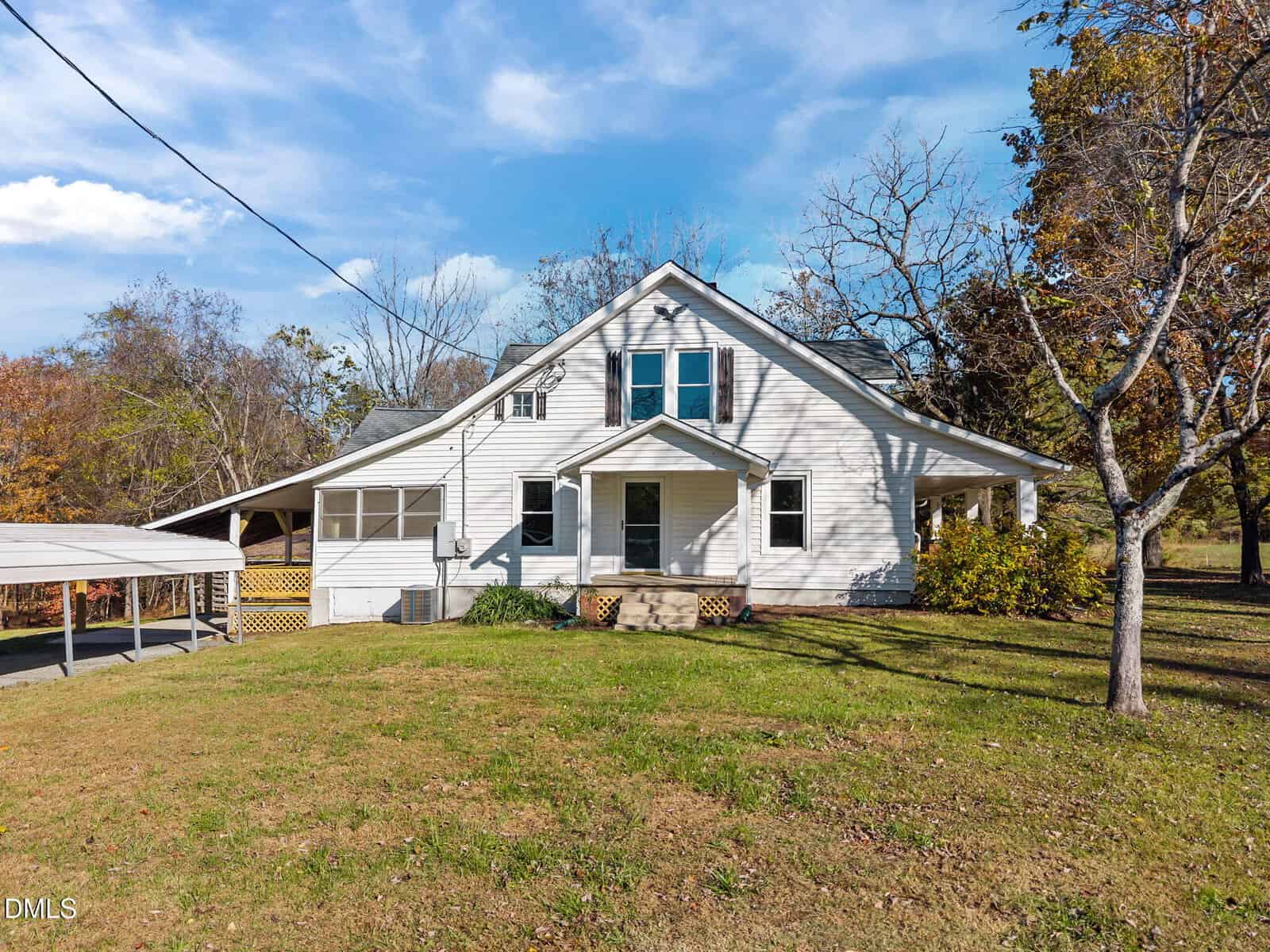 A white, two-story house with a covered porch sits on a grassy lawn. There are several windows, a carport on the left, and leafless trees surrounding the property. The sky is partly cloudy.