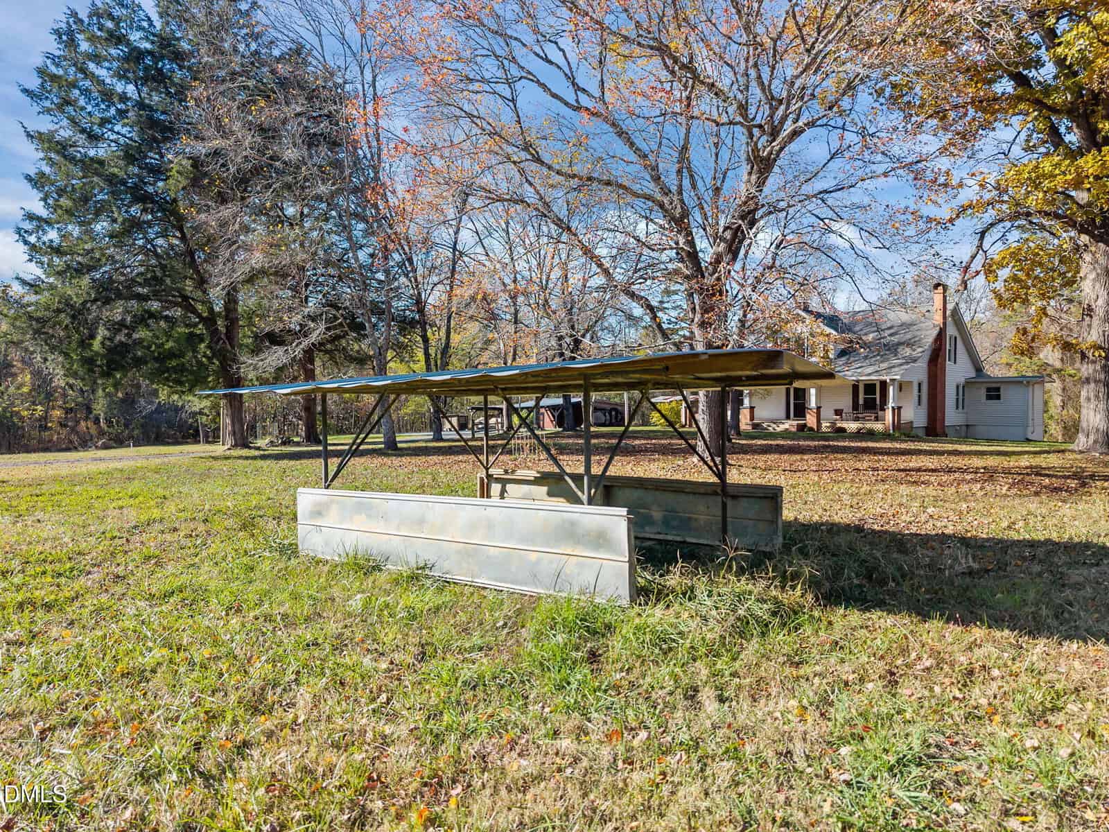 A metal livestock feeder sits on grassy ground in a yard, with mature trees and a white house with a porch and chimney visible in the background under a clear sky.