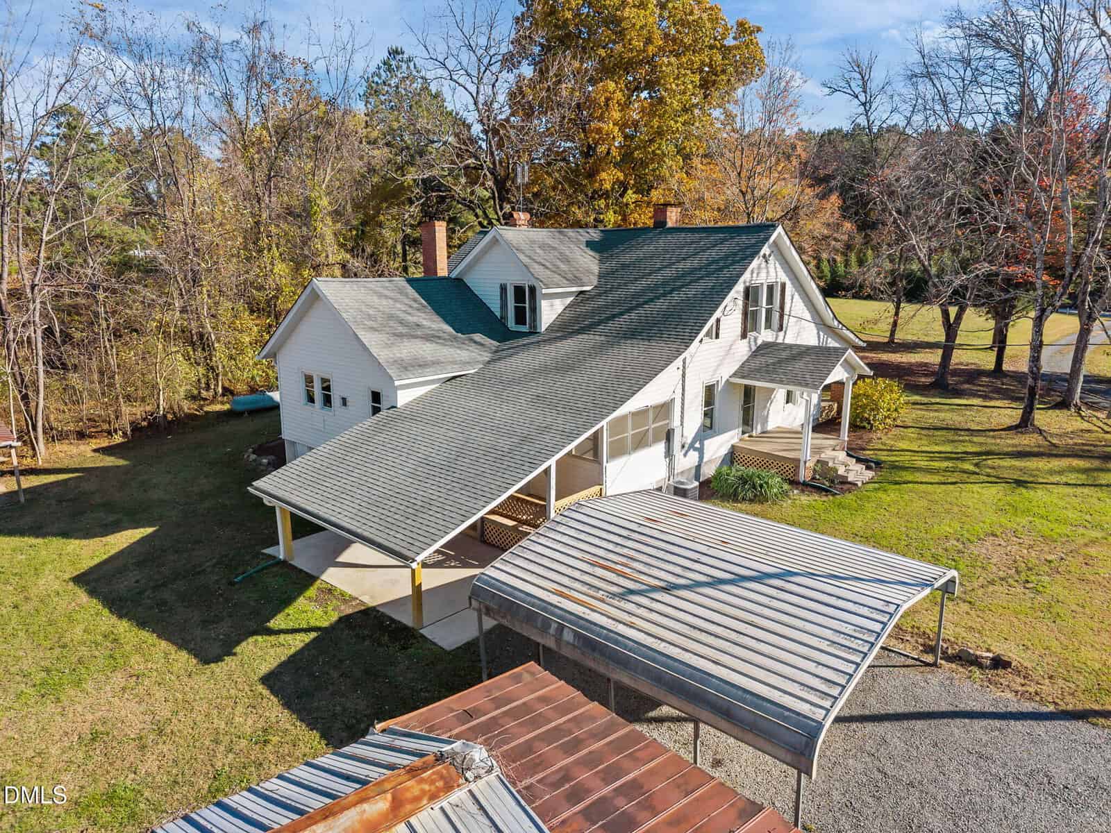 A large white house with a green gabled roof, a covered porch, and a metal carport sits on a grassy lot surrounded by trees with autumn foliage and a gravel driveway.