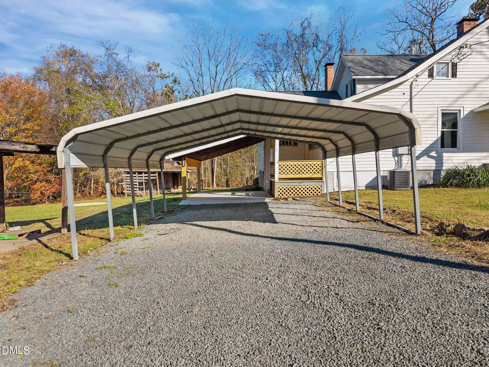 A metal carport with a gravel driveway underneath is positioned next to a white house with siding. Trees with autumn leaves are visible in the background.