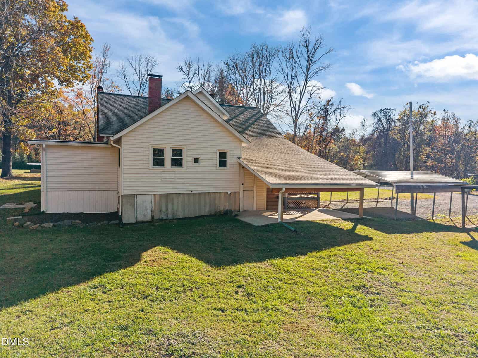 A beige house with a sloped roof, attached carport, concrete foundation, and grassy yard, photographed on a sunny day with trees and a blue sky in the background.